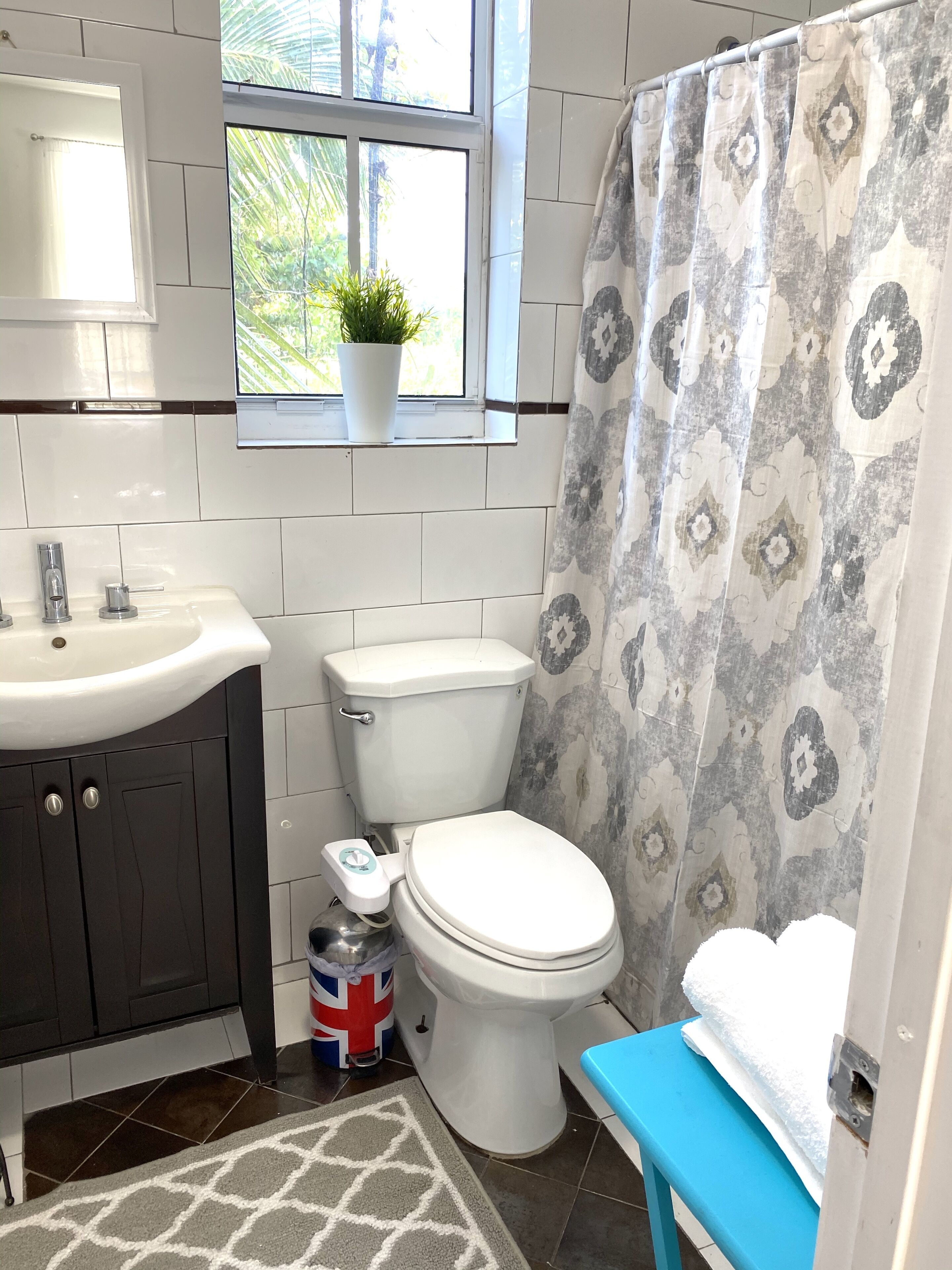 A bathroom with a white sink, a toilet, a blue bench, a window with a plant, and a shower curtain with a geometric pattern.
