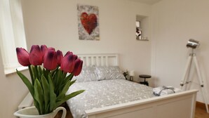 A bedroom with a bed, a white nightstand, a white vase with flowers, a camera on a tripod, and a heart-shaped painting on the wall.