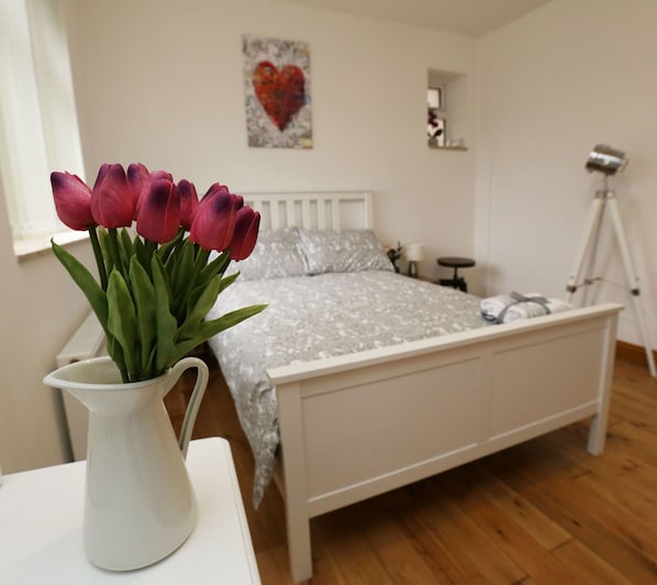 A bedroom with a bed, a white nightstand, a white vase with flowers, a camera on a tripod, and a heart-shaped painting on the wall.