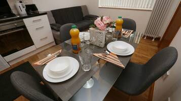 A dining area with a glass table set for four, including plates, glasses, and cutlery. A coffee maker and a vase with flowers are on the table.
