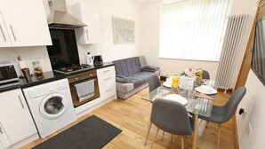 A compact kitchen with a washing machine, a dining area with a glass table, and a grey sofa.