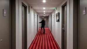 Een hotelgang met een rode loper, deuren aan weerszijden en een persoon die de gang in loopt.