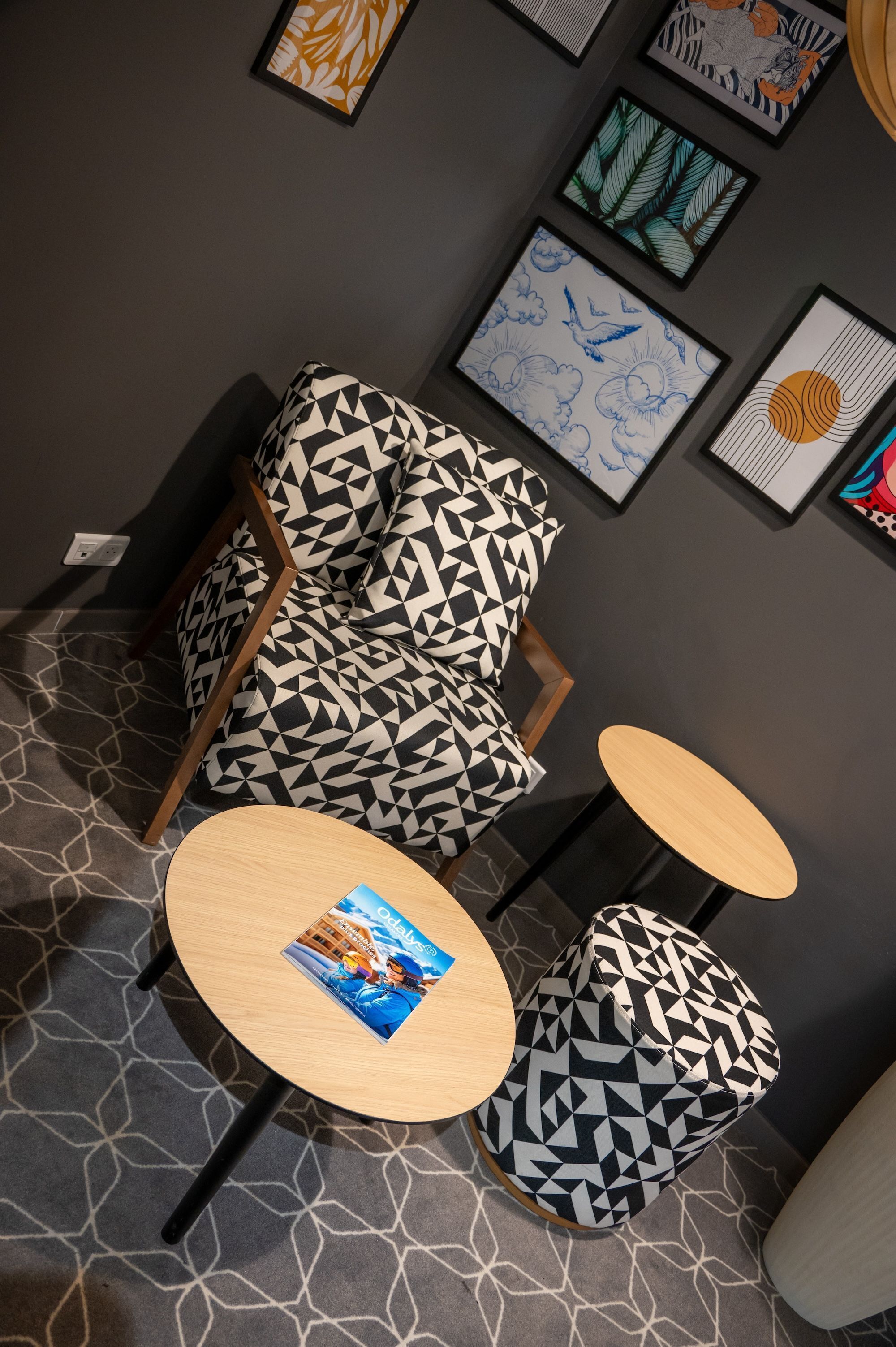 A modern lounge area with geometric patterned chairs and tables, framed artwork on the walls, and a patterned carpet.