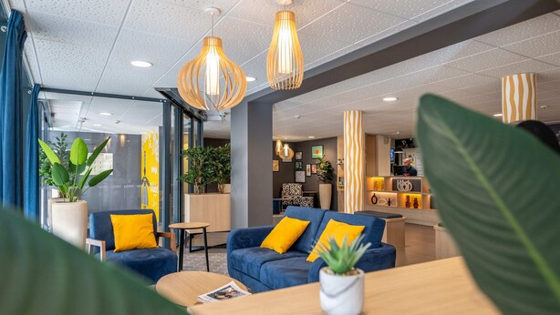 Een moderne hotellobby met een blauwe bank, gele kussens en hangende吊り下げ型lichtarmaturen.