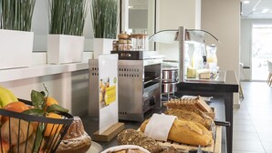 Zona de bufé con panes y pasteles variados, una cesta de frutas y un dispensador de zumos.
