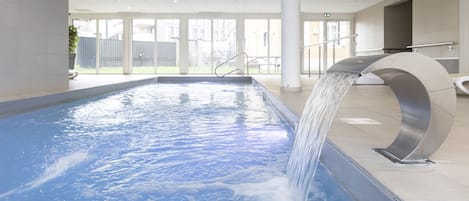 A swimming pool with a water feature, indoor lighting, and a view of the outdoors through large windows.
