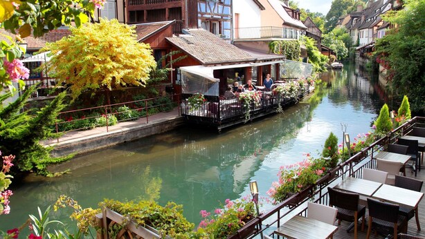 A riverside restaurant with outdoor seating overlooking a canal, surrounded by traditional European architecture.