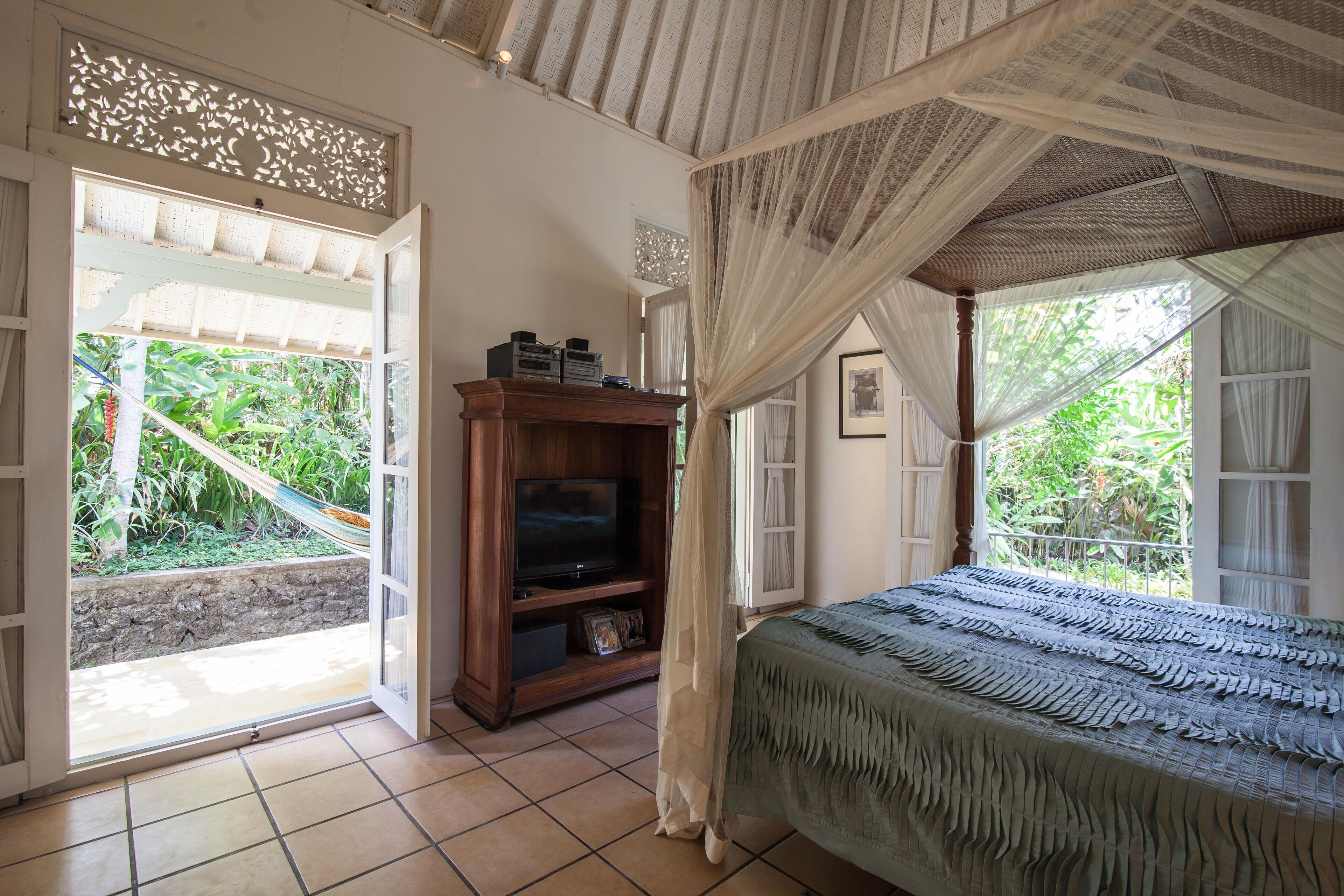 Quarto com cama, televisão sobre suporte de madeira e vista para o verde lá fora.