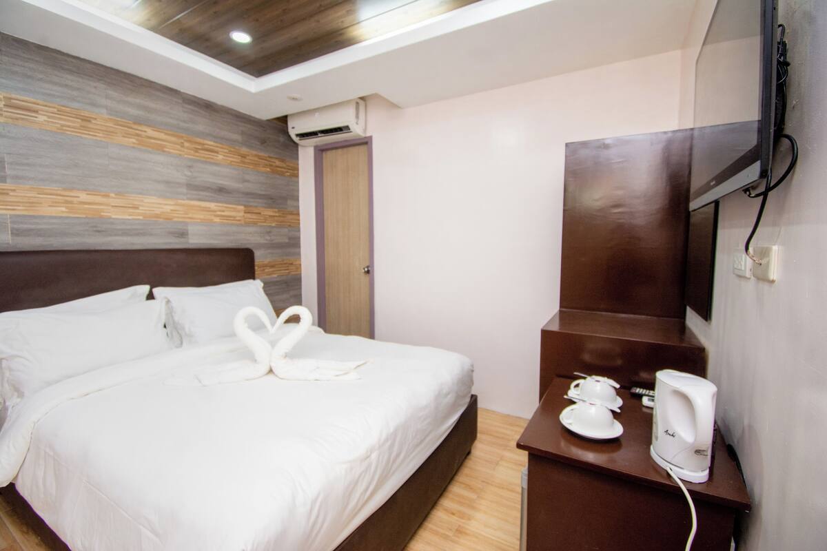 Quarto de hotel com cama, cabeceira de madeira, televisão de ecrã plano na parede e uma pequena mesa com chaleira e chávenas.
