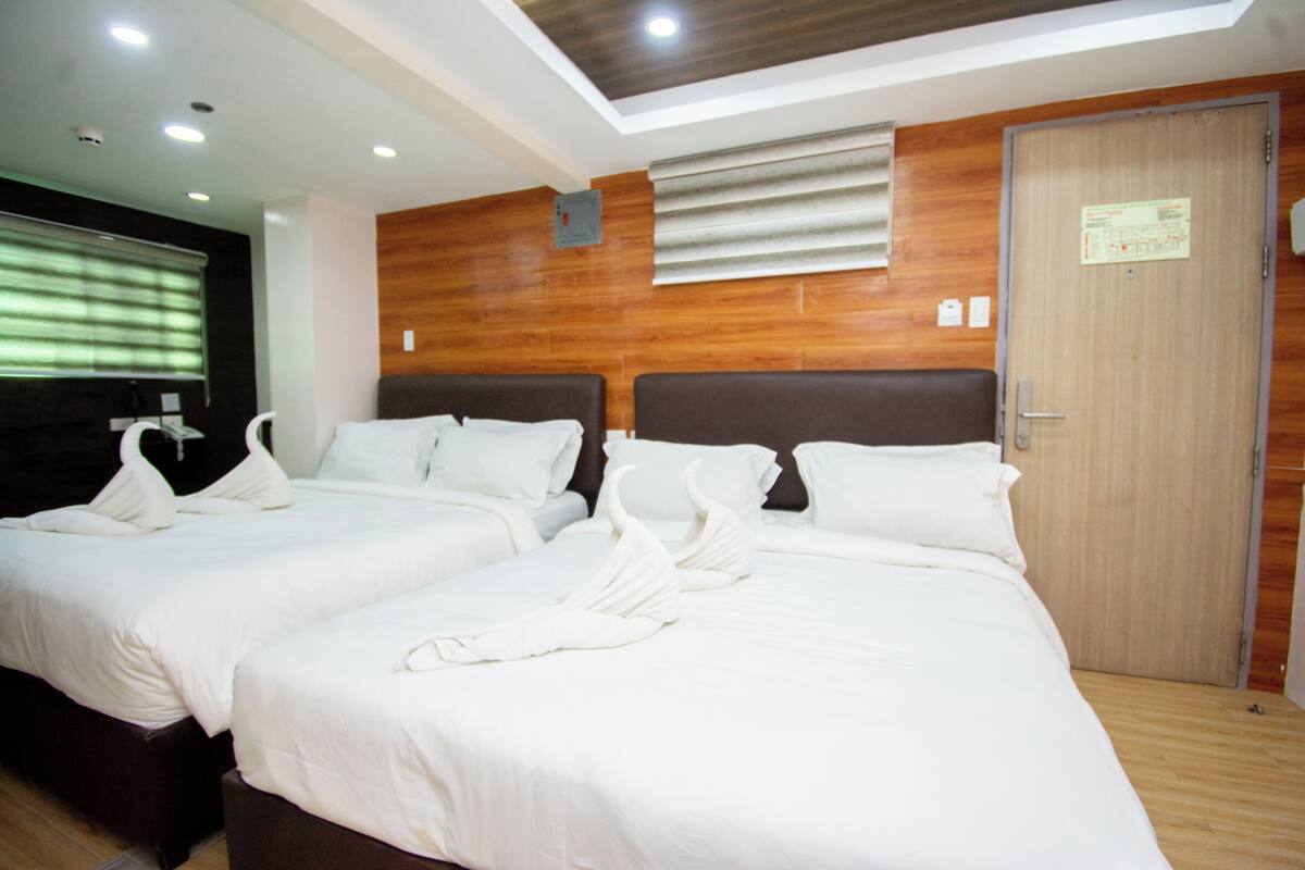 Quarto de hotel com duas camas, painéis de madeira nas paredes, teto com iluminação embutida e uma porta com uma placa.