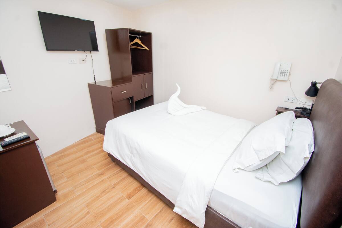 Quarto de hotel com uma cama, guarda-roupa, escrivaninha, televisão e telefone.