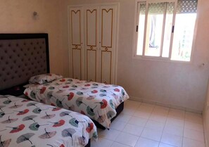 A bedroom with two beds, a tufted headboard, a window with blinds, and a door with decorative panels. - Residence Atlas Marrakech 4677 (Marrakech)