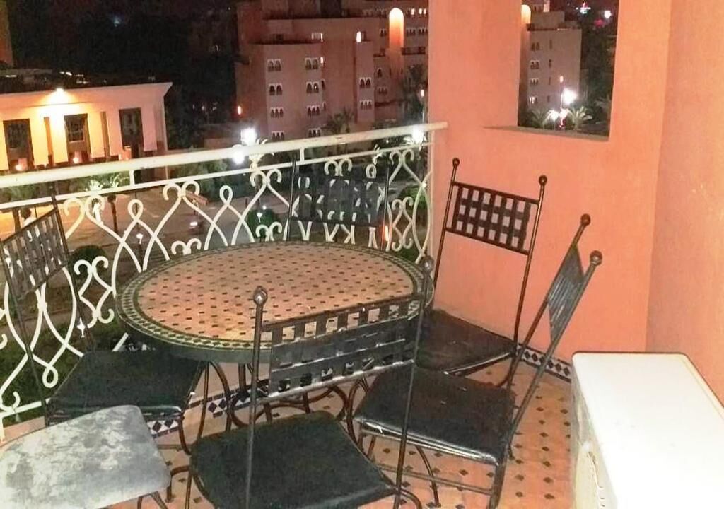 Un balcon avec une table ronde et des chaises, un mur blanc et une vue sur les bâtiments la nuit.
