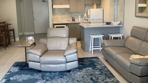 Una sala de estar moderna con un sofá de cuero, un comedor con una mesa pequeña y sillas, y una cocina con armarios grises y un refrigerador blanco.