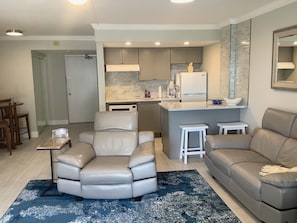 A modern living room with a leather sofa, a dining area with a small table and chairs, and a kitchen with grey cabinets and a white refrigerator.