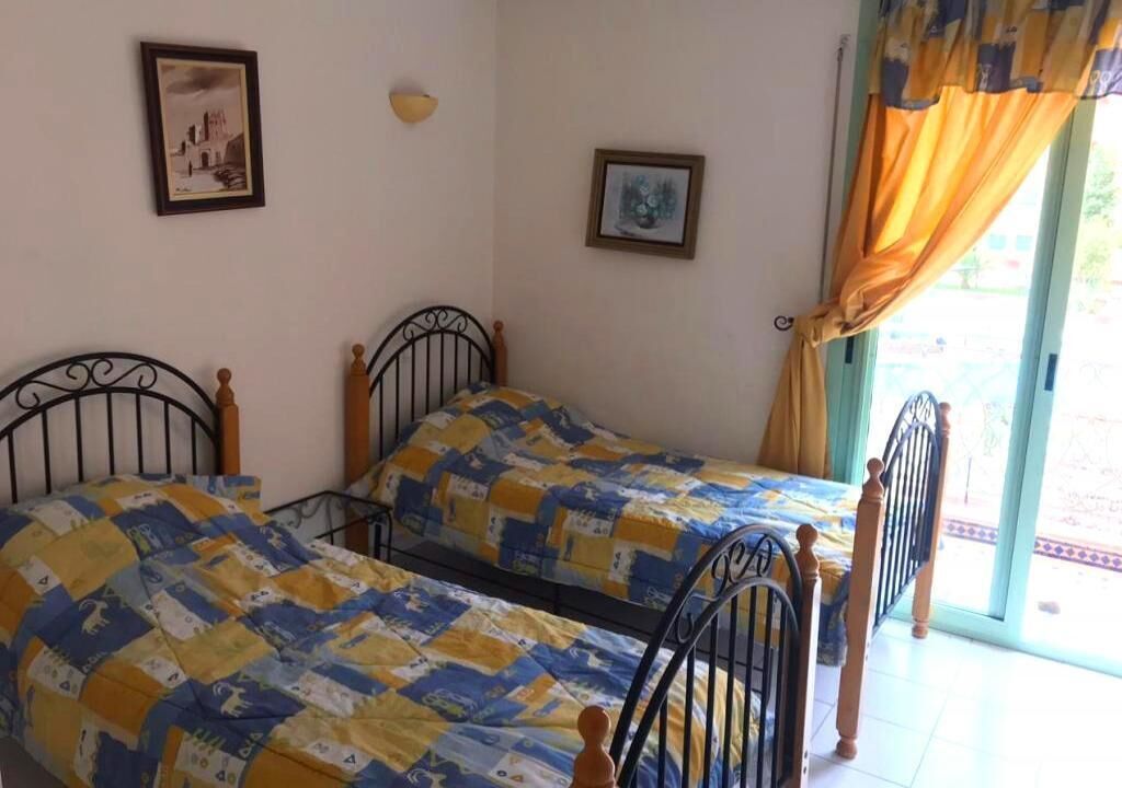 Una habitación con dos camas, cada una con cabecera de madera y colcha estampada. Hay dos cuadros enmarcados en la pared, un aplique y una ventana con cortinas.