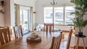 Uma sala de jantar com mesa e cadeiras de madeira, um candelabro e vista para a água através de grandes janelas.