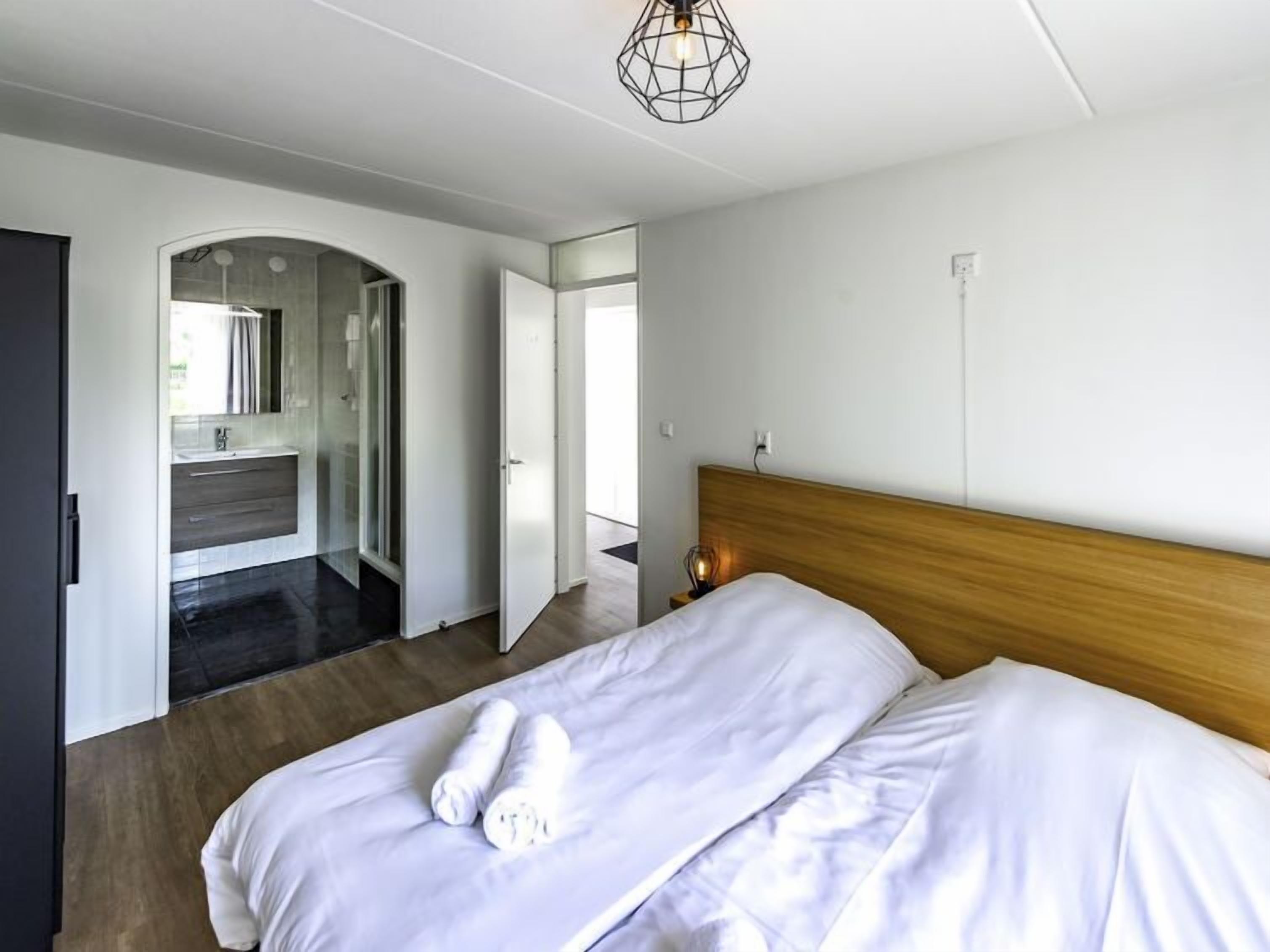 A modern hotel room with a wooden headboard, a large bed with white linens, a nightstand with a lamp, and a bathroom visible through an archway.