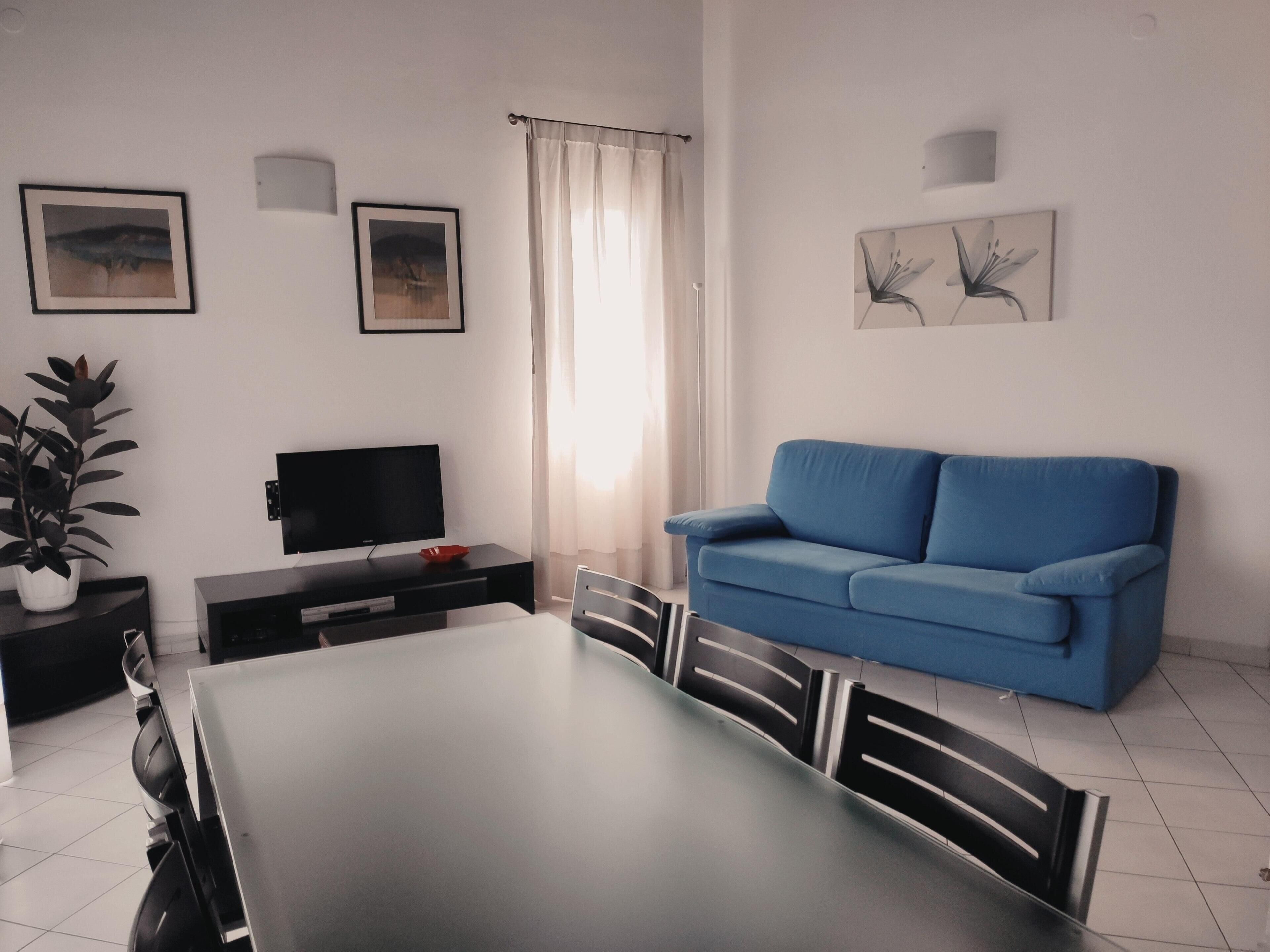 Un salon moderne avec un canapé bleu, un téléviseur à écran plat, une table à manger avec des chaises et des œuvres d’art accrochées aux murs.