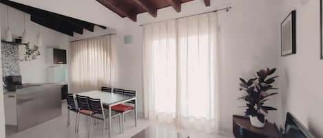 Una cocina moderna con armarios blancos, un comedor con mesa de vidrio y una sala de estar con televisor y planta.