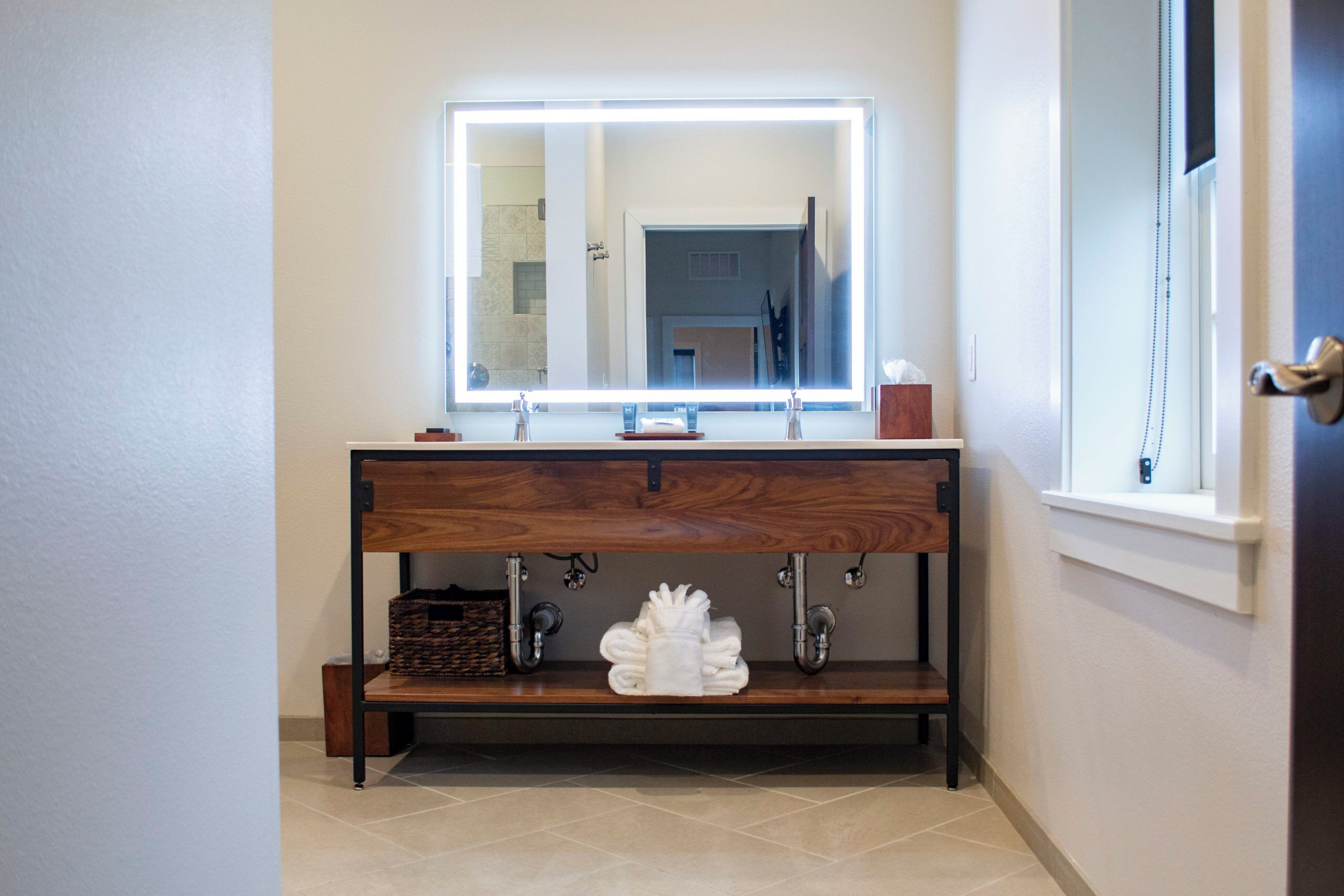 A bathroom with a wooden vanity, a large mirror with built-in lighting, and a window.