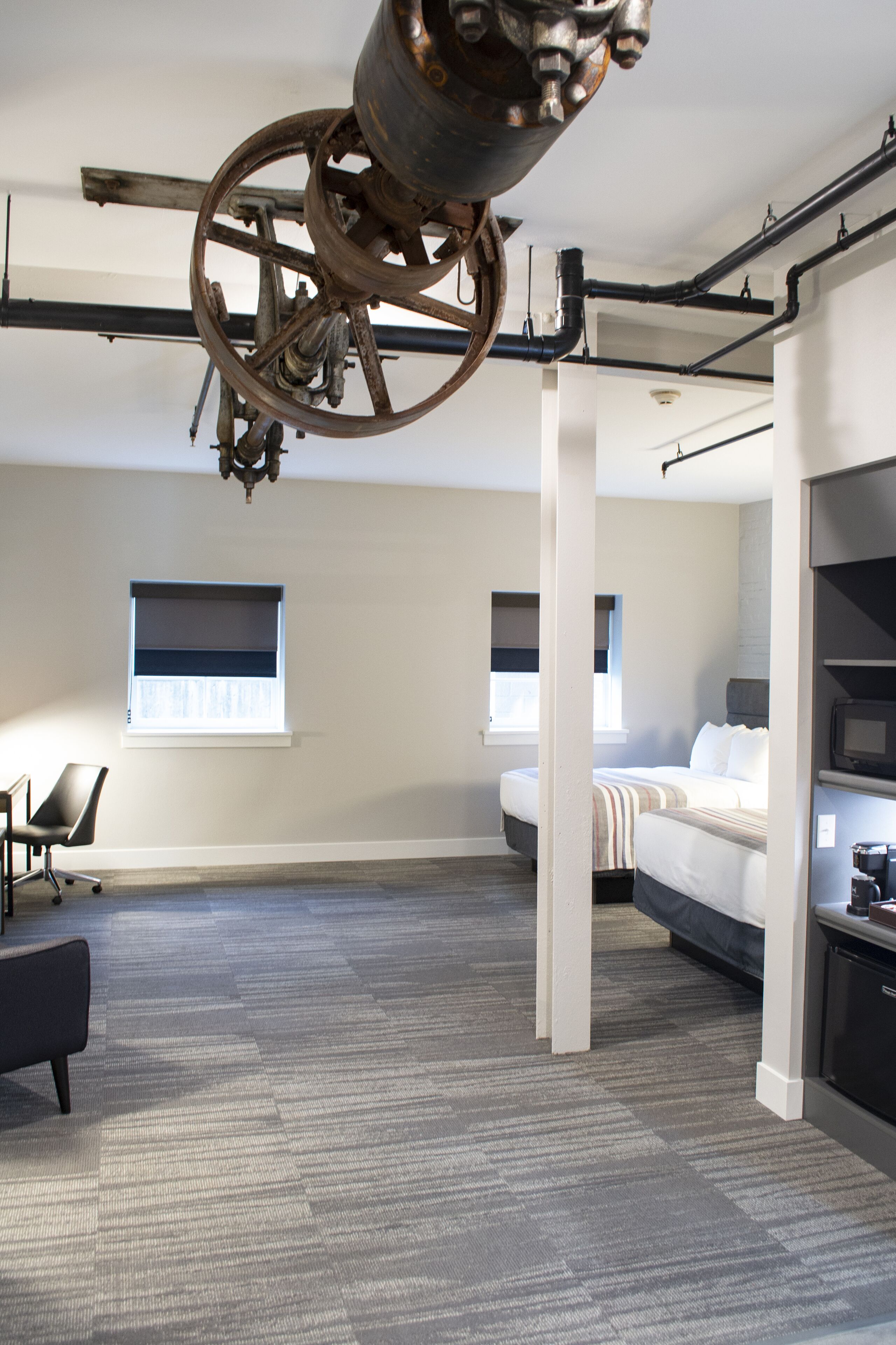 Una habitación de hotel moderna con una cama grande, un escritorio y una silla. Hay dos ventanas y un techo con tuberías a la vista y una gran rueda de metal.