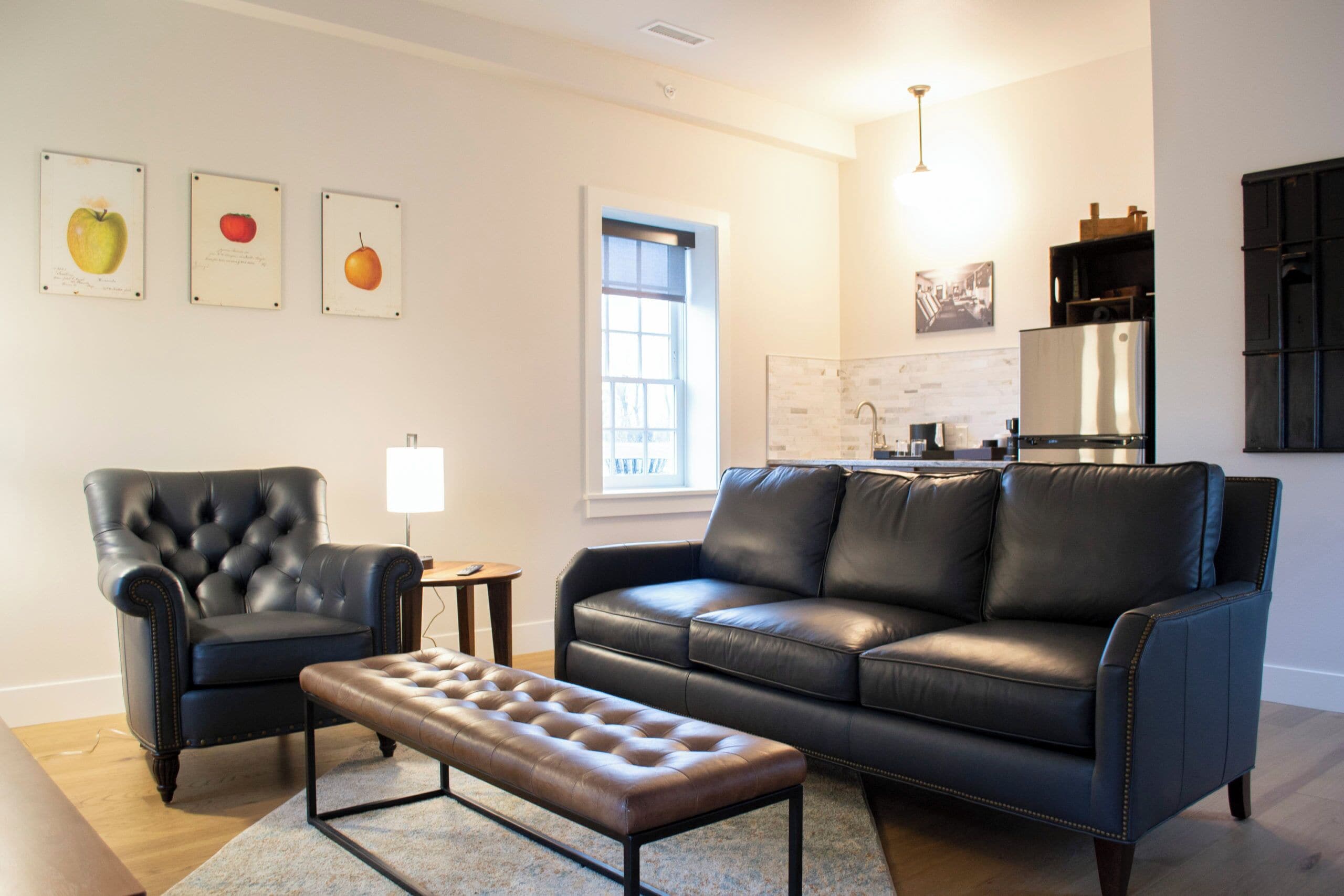 A modern living room with a leather sofa, a tufted ottoman, and a black leather armchair. There is a window with a view of the outside, a kitchen area with a refrigerator, and a wall-mounted artwork of fruits.