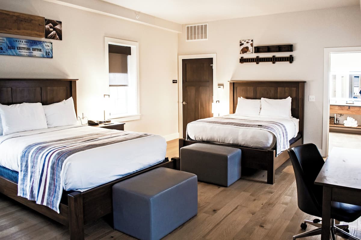 Une chambre d’hôtel avec deux lits, un bureau et une chaise. On y trouve également deux poufs, une fenêtre et une porte.