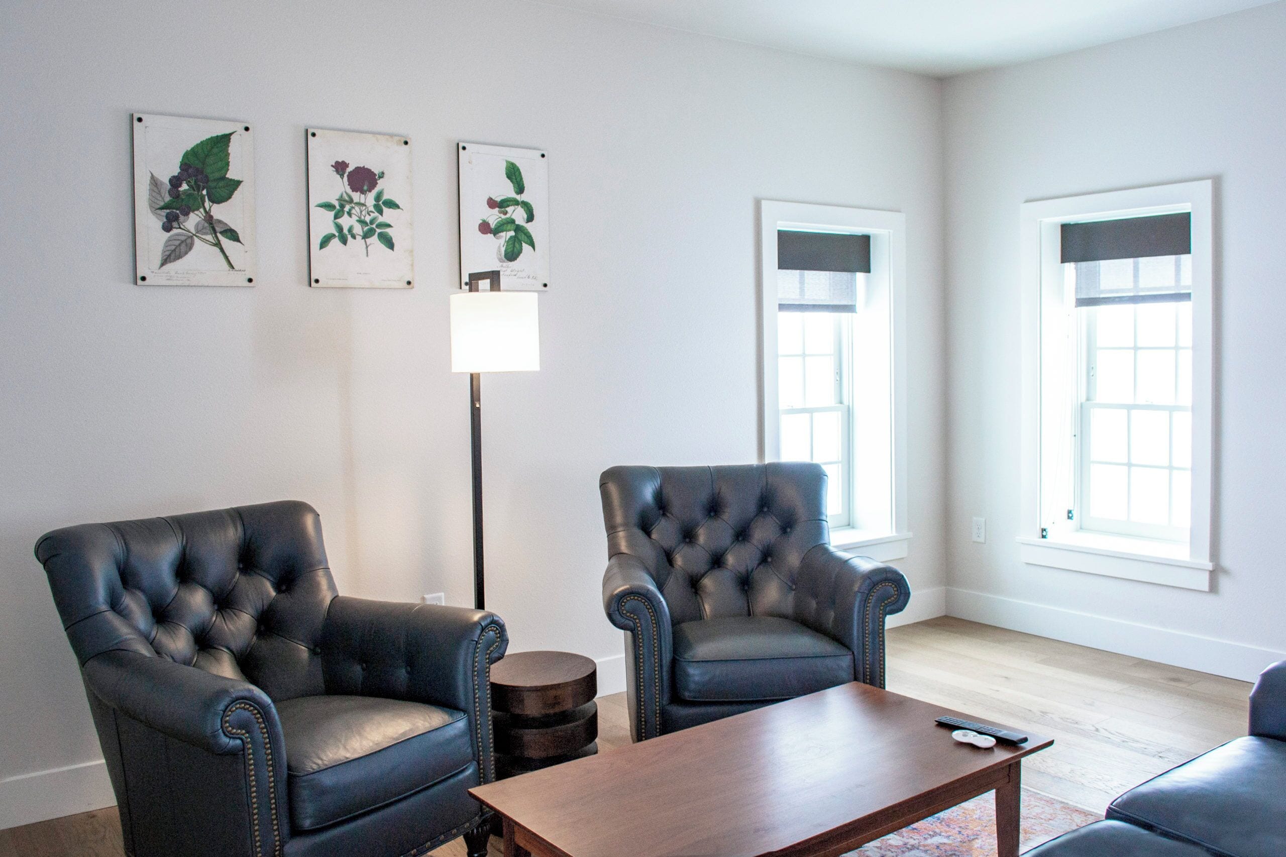 A living room with two black leather armchairs, a wooden coffee table, and three framed botanical prints on the wall.