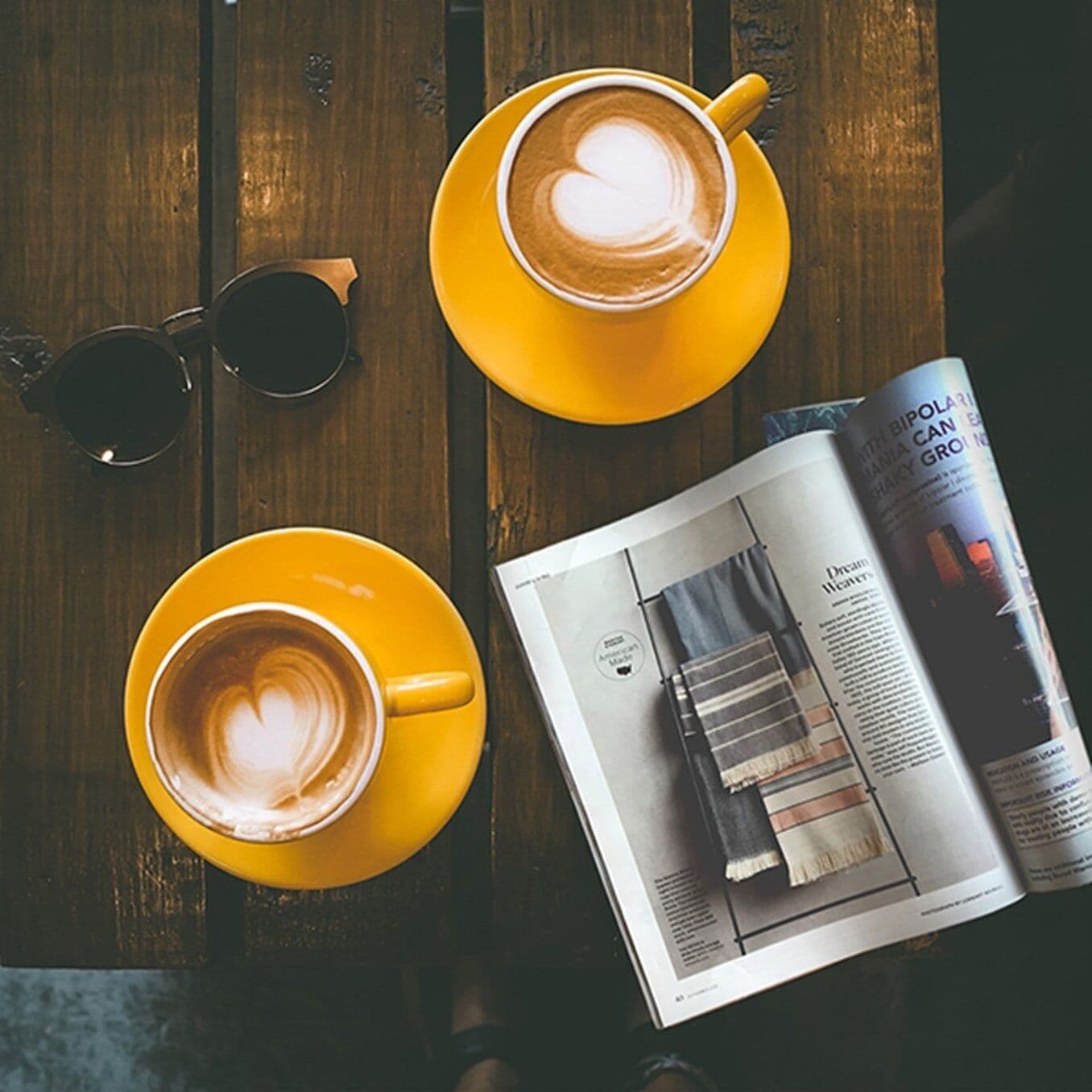 Dos tazas de café amarillas sobre una mesa de madera, un par de gafas de sol y una revista con publicidad de café.