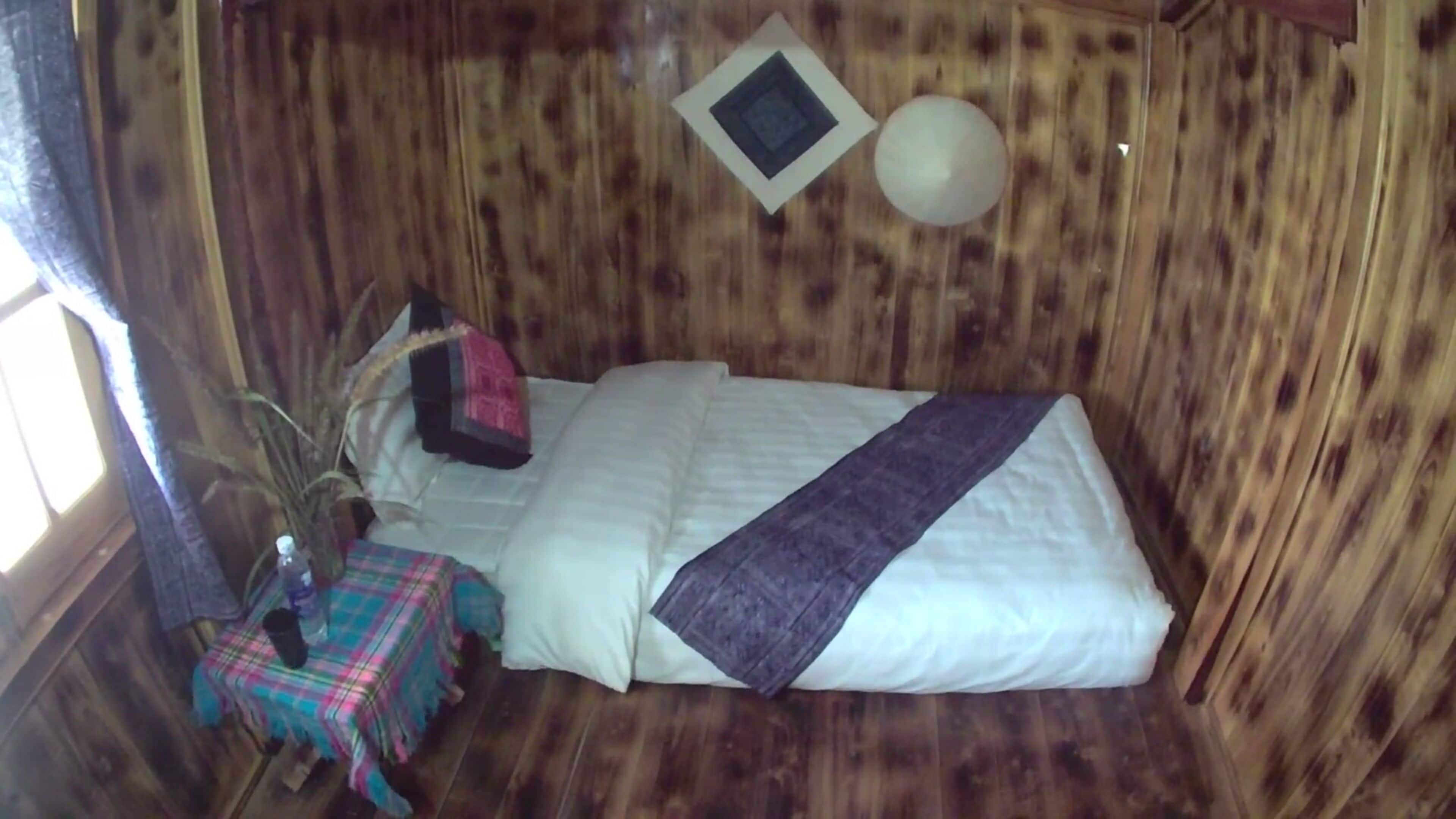 A small, cozy room with a single bed, a small table with a colorful tablecloth, and a potted plant.