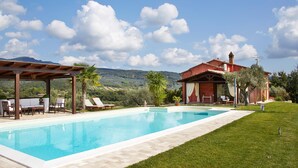 Une piscine avec des chaises longues et une pergola, un bâtiment rouge doté d’une cheminée, et un paysage montagneux en arrière-plan.
