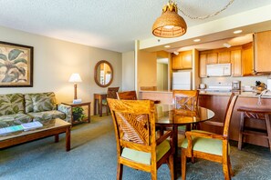 A living room with a sofa, coffee table, and dining table with chairs. There is a kitchen area with cabinets and appliances. - Aston Ka'anapali Shores 550 | Garden View Retreat – Pool, Hot Tub & Spa (Lahaina)