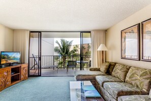 A hotel room with a balcony, a sofa, a TV on a wooden cabinet, and a table with a lamp.