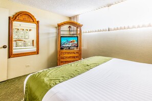 A hotel room with a bed, a television on a wooden stand, a mirror, and a wall with a decorative tile pattern. - Aston Ka'anapali Shores 550 | Garden View Retreat – Pool, Hot Tub & Spa (Lahaina)