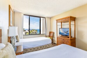 Habitación de hotel con dos camas, un televisor sobre un mueble de madera y un amplio ventanal con vistas a palmeras y al océano.