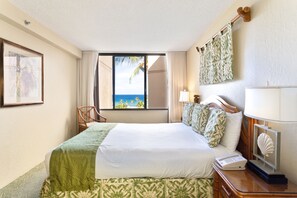 Un cuarto de hotel con una cama grande, vista al océano, una silla de madera, un escritorio con lámpara y un cuadro en la pared.