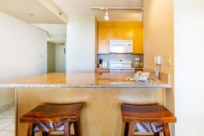A kitchen with a granite countertop, wooden stools, a microwave, and a coffee maker. - Sweeping Ocean Views | Aston Ka'anapali Shores 727 – Pool, Hot Tub & Spa (Lahaina)