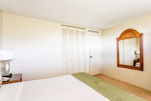 Quarto de hotel com cama, mesa de cabeceira com abajur, espelho, armário com portas venezianas e piso coberto por carpete.