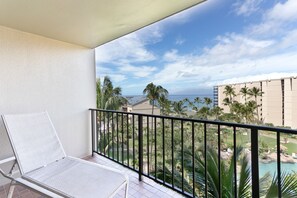 Ein Balkon mit weißem Stuhl, Blick auf einen Strand, Palmen und ein Gebäude im Hintergrund.