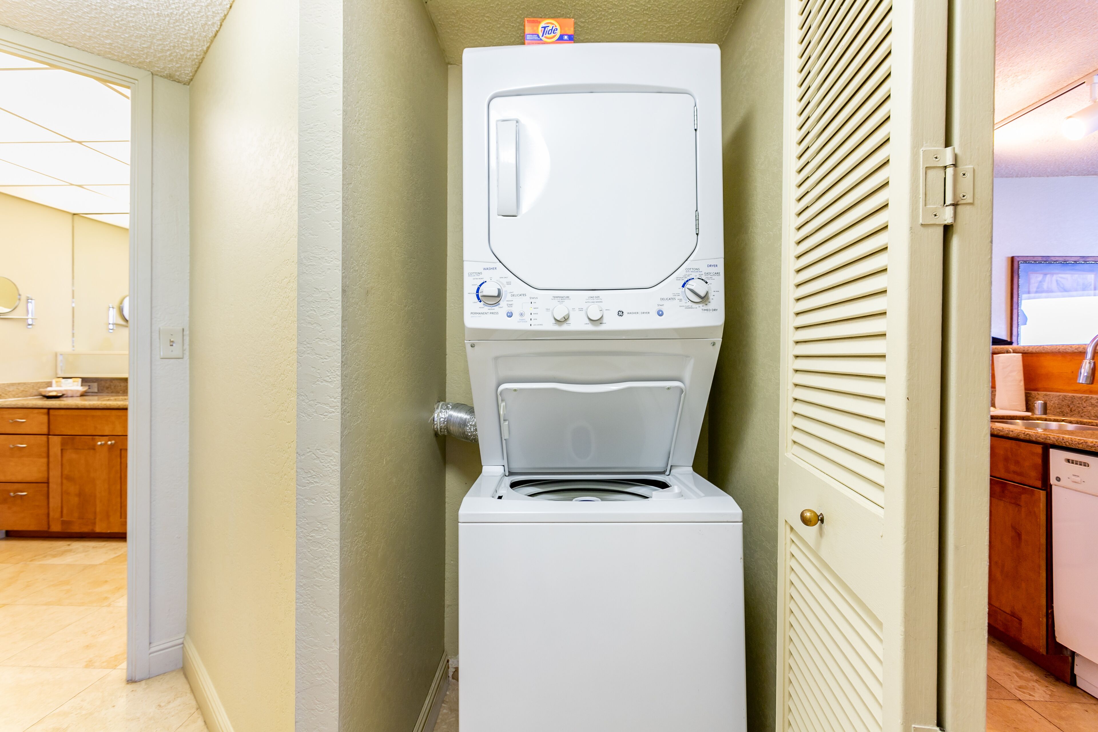 Un lave-linge et un sèche-linge compacts sont installés dans un couloir, avec un meuble en bois et une partie de la cuisine visible au fond.