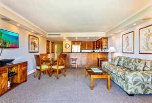 Un salon comprenant un canapé, une table basse et des fauteuils, un téléviseur, une horloge, ainsi qu’un coin cuisine avec des placards et un réfrigérateur.