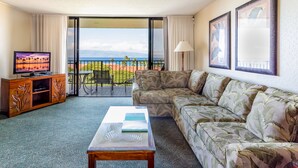 Ein Hotelzimmer mit einer Couch, einem Couchtisch, einem Fernseher auf einem Holzschrank und einem Balkon mit Blick auf Strand und Berge.