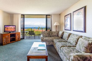 Une chambre d’hôtel avec un canapé, une table basse, un téléviseur posé sur un meuble en bois et un balcon offrant une vue sur la plage et les montagnes.