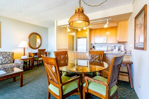 A dining area with a round glass table and wooden chairs, a kitchen with wooden cabinets, and a living room with a patterned sofa and a lamp.