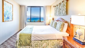Habitación de hotel con una cama grande, vista al océano, cabecera de madera, mesita de noche con lámpara y una concha decorativa.