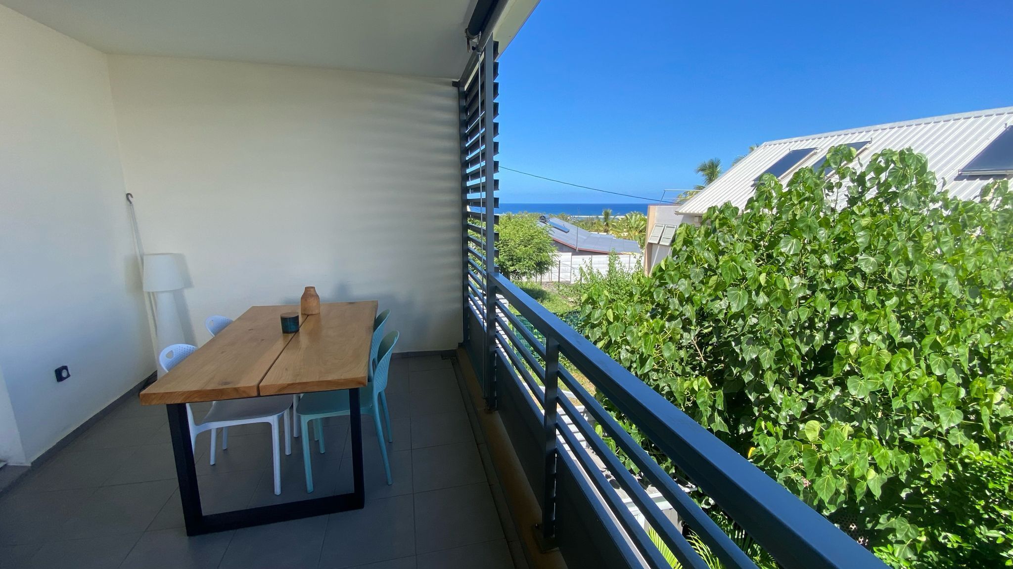 En balkon med et træbord og stole, havudsigt og en hæk i baggrunden.