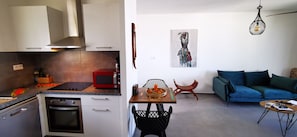 A modern kitchen with a microwave, oven, and stainless steel appliances. There is a dining area with a table, chairs, and a blue sofa. A painting is on the wall, and a hanging light fixture is visible. - Appart "Rev'Alizé" in the heart of the Saline lagoon (La Saline-Les-Bains)