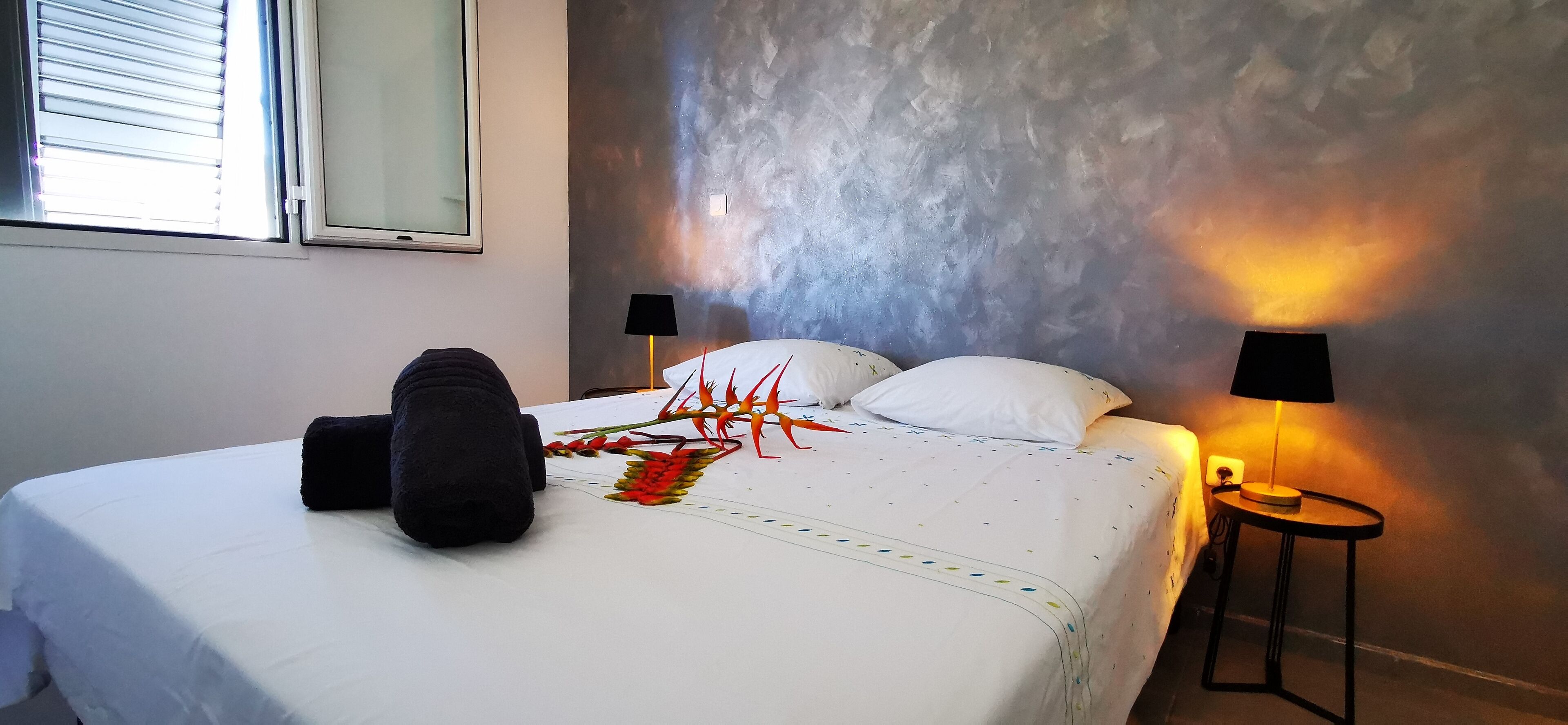 A neatly made bed with a white comforter and decorative pillows, a black throw blanket, and a colorful decorative piece. There are two bedside lamps on either side of the bed, and a small table with a lamp next to the bed.