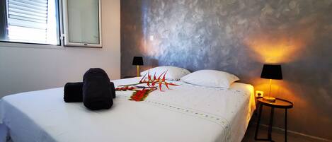 A neatly made bed with a white comforter and decorative pillows, a black throw blanket, and a colorful decorative piece. There are two bedside lamps on either side of the bed, and a small table with a lamp next to the bed.
